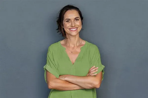 Hormones-for-Women-Doctor A woman in a green shirt stands smiling against a gray-blue wall, pleased with her hormone therapy from Christine Collins, MD in Westside/Beverly Hills.