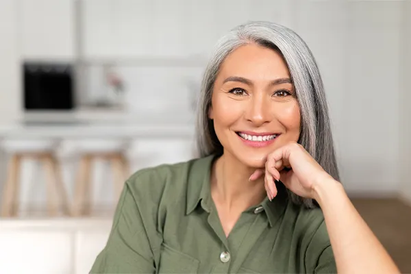 Menopause-Clinic An asian woman with long gray hair and a green shirt sitting and smiling on her couch, happy with her menopause care from [NAME} in Westside/Beverly Hills.
