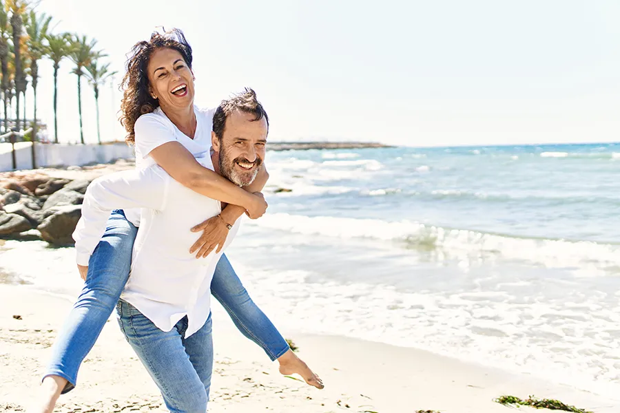 NAD-Therapy-Clinic A woman piggybacks on her husband as the middle-aged couple have fun at the beach. Get NAD Therapy from Christine Collins, MD in Westside/Beverly Hills.