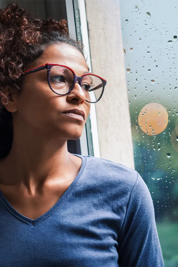 PMDD-Treatment A black woman with with a somber expression and glasses and a gray shirt stares out the window on a rainy day. Get treatment for Premenstrual Dysphoric Disorder from Christine Collins, MD in Westside/Beverly Hills.