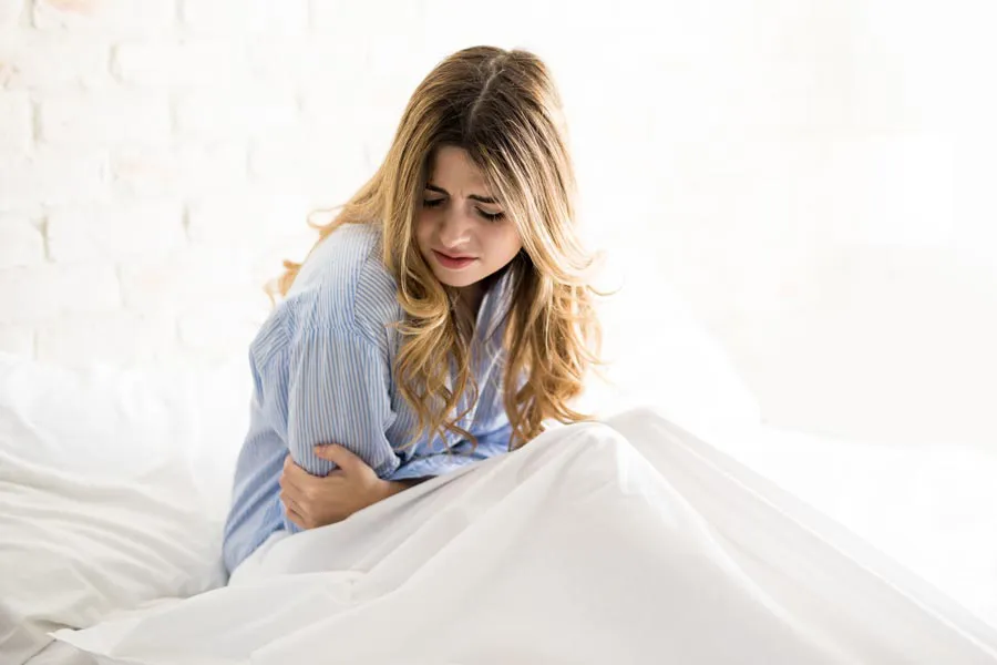 PMS-Therapy A woman with long blonde hair sits in her bed holding her stomach in pain, considering getting care for PMS from Christine Collins, MD in Westside/Beverly Hills.