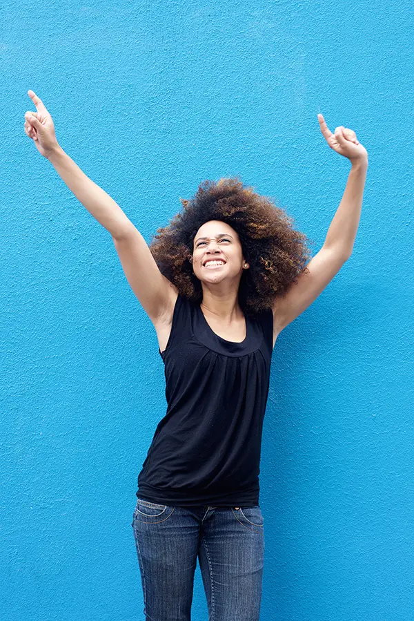 PMS-Treatment A woman in a dark blue tank top standing in front of a bright blue wall, raising her arms in celebration of relief from PMS from Christine Collins, MD in Westside/Beverly Hills.
