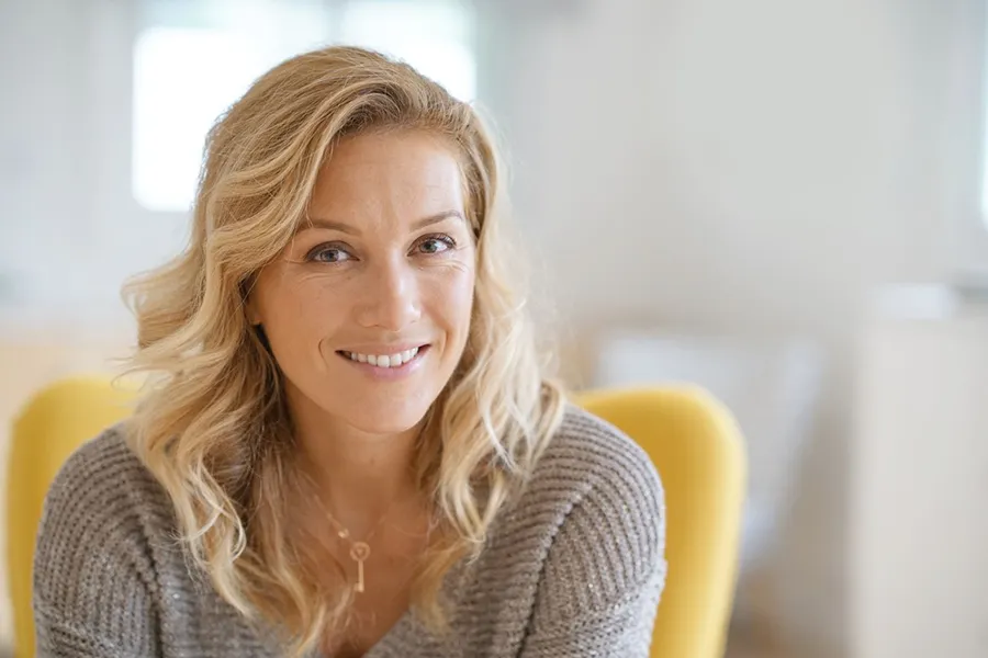 Perimenopause-Doctor A blonde woman wearing a a gray sweater sits inside in a yellow chair, smiling and happy with her perimenopause treatment from Christine Collins, MD in Westside/Beverly Hills.