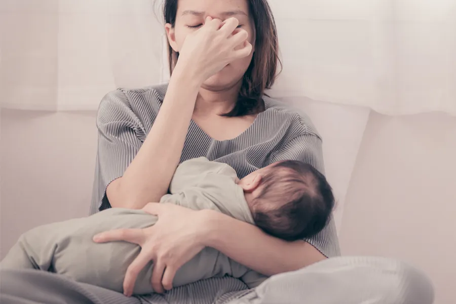 Postpartum-Hormone-Imbalance-Clinic A woman nursing an infant on couch rubs her eyes with exhaustion. Get treatment for postpartum hormone imbalance from Christine Collins, MD in Westside/Beverly Hills.